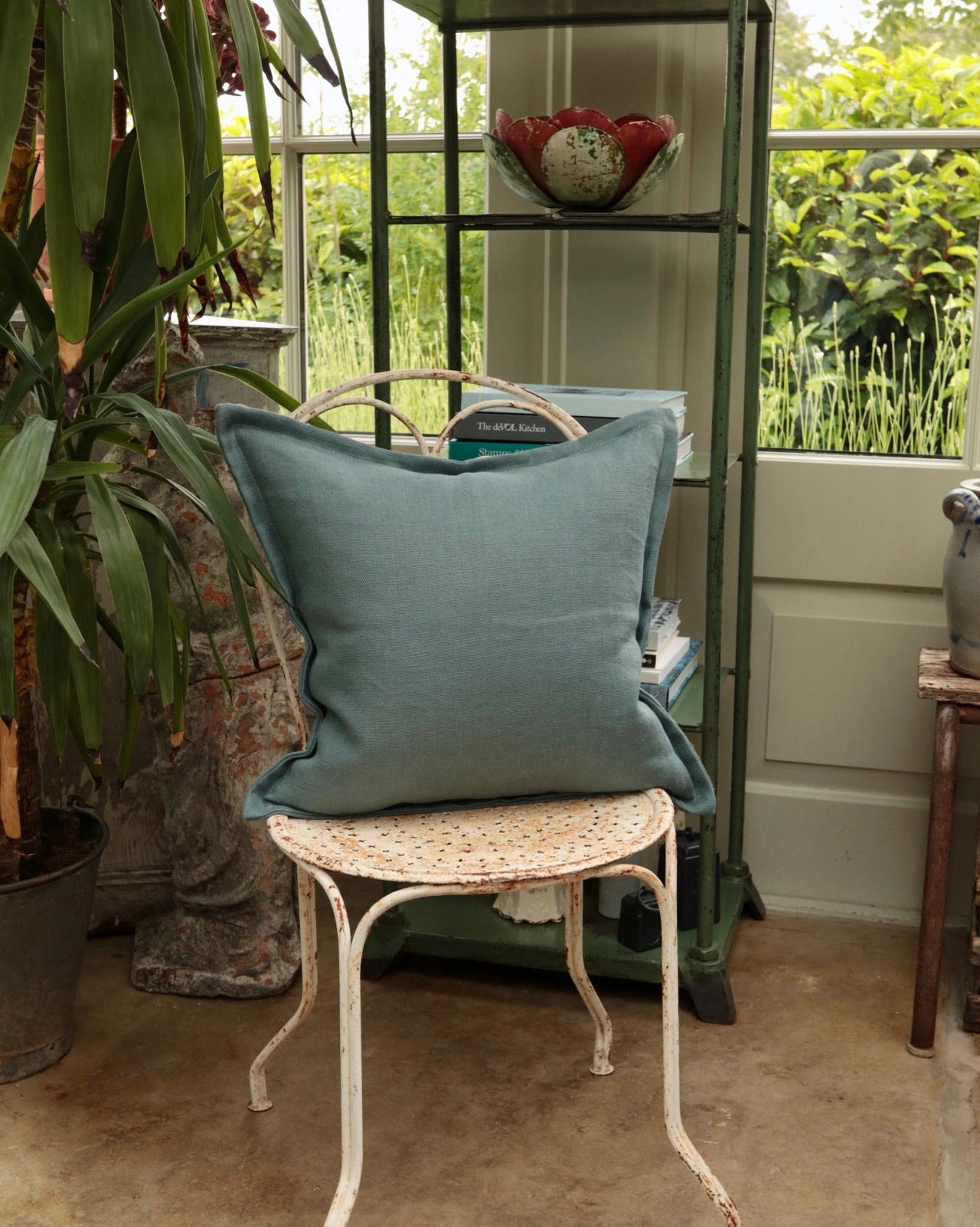 Blue cushion on a white metal chair with a green shelf and plants in the background