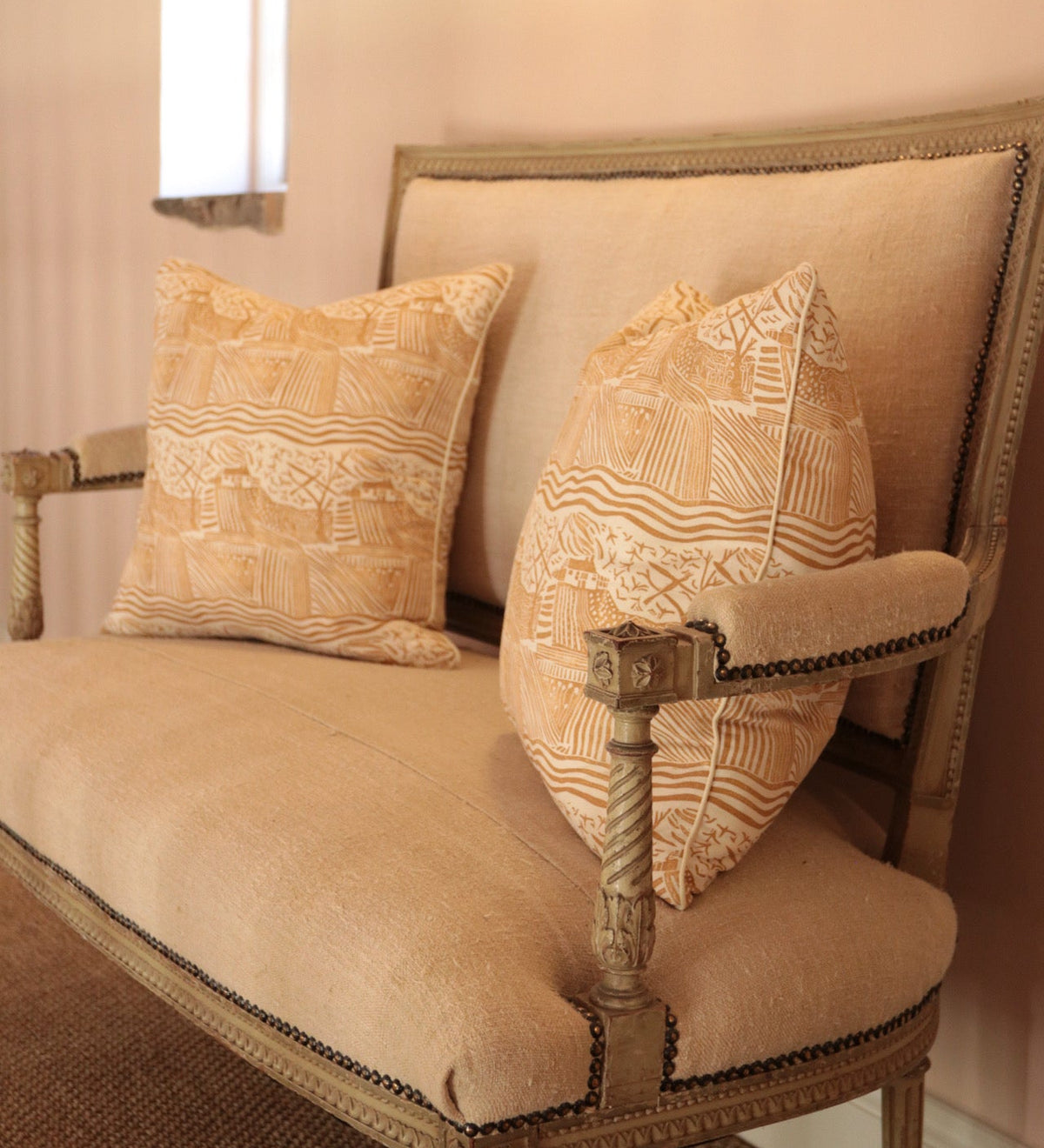 A beige upholstered armchair with a distressed wood frame and two matching patterned cushions, styled in a warm, softly lit interior setting.